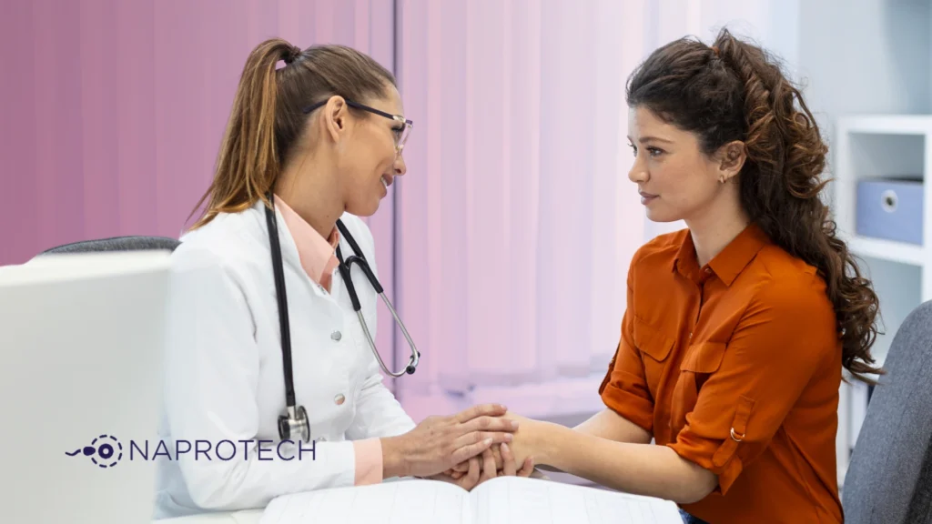Doctora conversa con una paciente en un consultorio médico. La profesional, con bata blanca y estetoscopio, sostiene las manos de la mujer en señal de apoyo. Fondo con tonos rosados y logo de Naprotech visible en la esquina inferior izquierda.