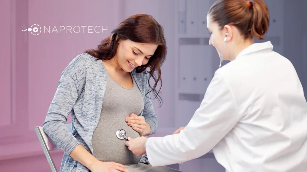 Médica revisando a una mujer embarazada con estetoscopio durante una consulta de control prenatal, con el logo de Naprotech al fondo