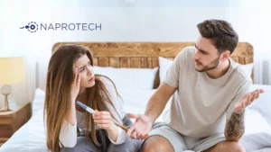 Pareja joven en una cama mostrando preocupación mientras observan una prueba de embarazo. La mujer sostiene el test con gesto de incertidumbre y el hombre parece frustrado. Imagen asociada al tema de fertilidad y diagnóstico natural con el logo de Naprotech