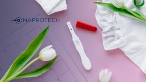 Prueba de embarazo con resultado positivo sobre una superficie rosada, acompañada de tulipanes blancos, un calendario y prendas de bebé, junto al logo de Naprotech.