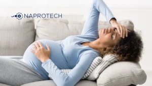 Mujer embarazada recostada en un sofá, con expresión de malestar mientras se toma la frente con una mano y sostiene su abdomen con la otra, junto al logo de Naprotech en la parte superior izquierda.