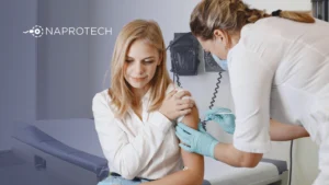 Médica aplica una inyección en el brazo de una mujer joven en un consultorio, mientras ella sostiene su brazo; al fondo se ve el logo de Naprotech.