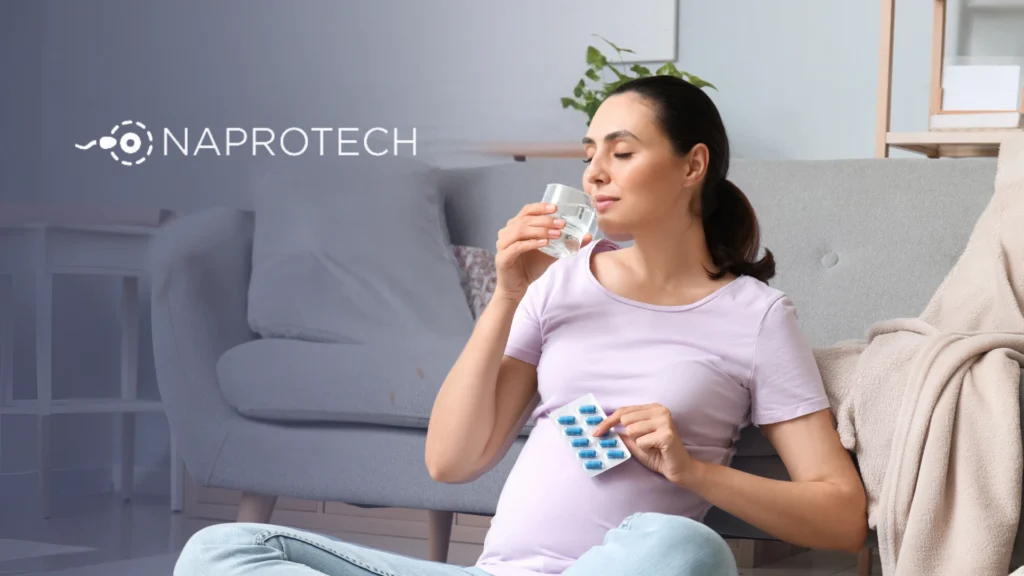 Mujer embarazada sentada en el suelo de una sala acogedora, sosteniendo un blíster de cápsulas azules mientras bebe un vaso de agua. A la izquierda se muestra el logo de Naprotech.