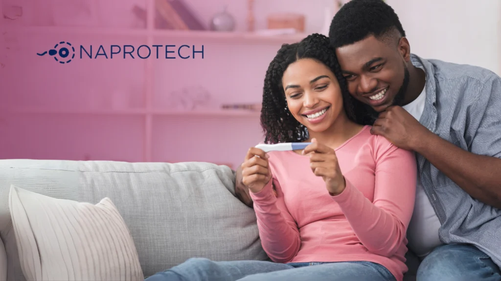 Pareja sentada en un sofá sonríe mientras observa una prueba de embarazo, con un fondo rosado suave y el logo de Naprotech al lado izquierdo.