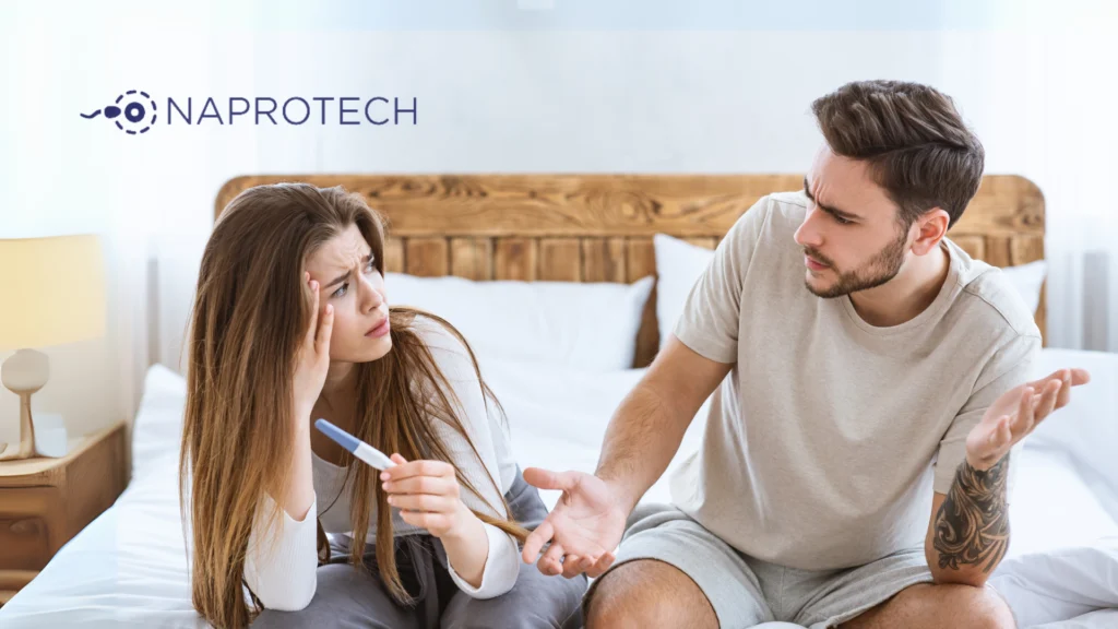 Pareja en la cama mirando una prueba de embarazo con preocupación, representando signos de posibles dificultades de fertilidad, junto al logo de Naprotech.